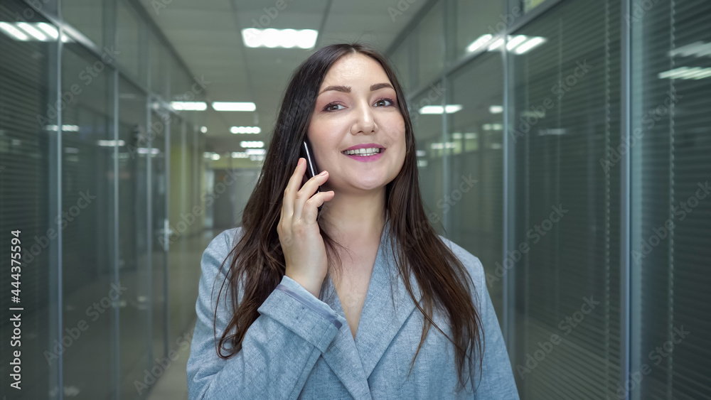 身穿灰色夹克的深色女商人站在大楼休息室里用黑色智能手机与魅力交谈