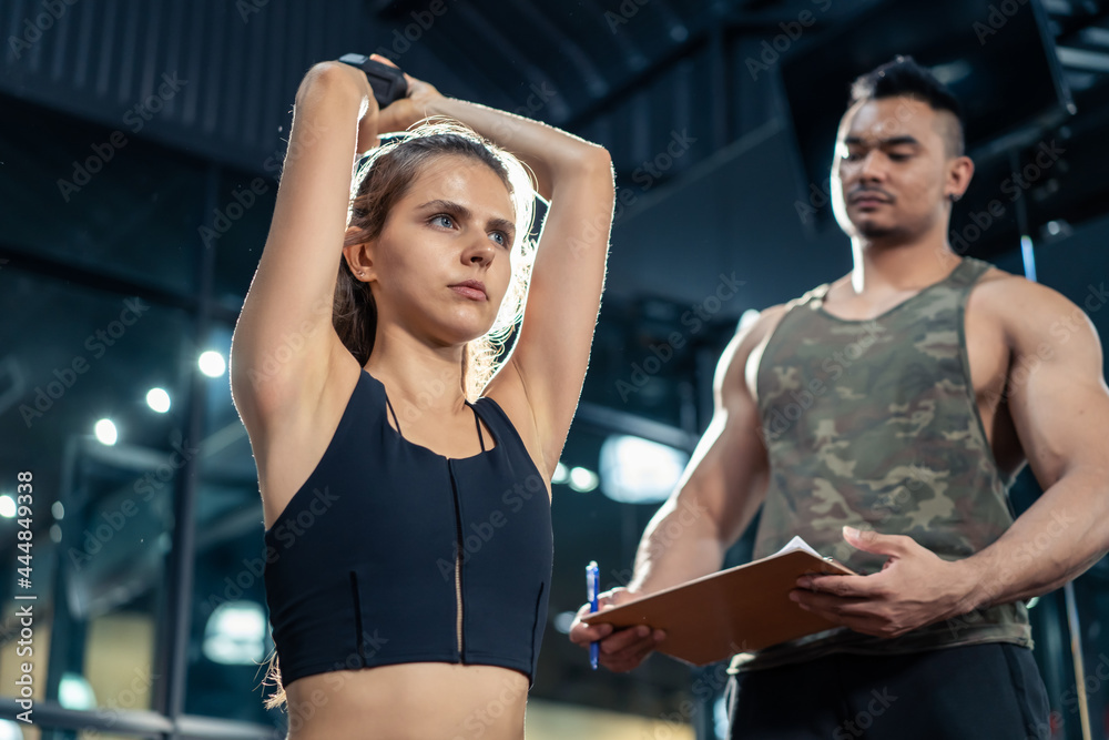 Asian trainer motivate young Caucasian lifting weight dumbbell in gym.