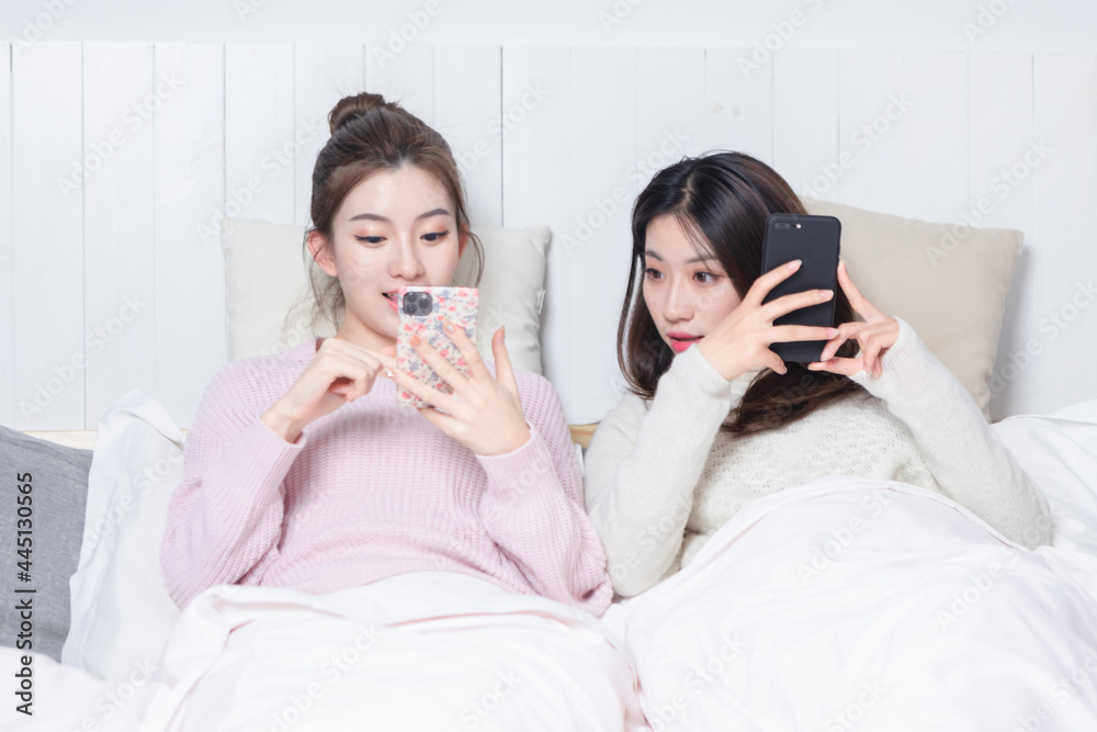 Two beautiful women lying on the bedside playing mobile games