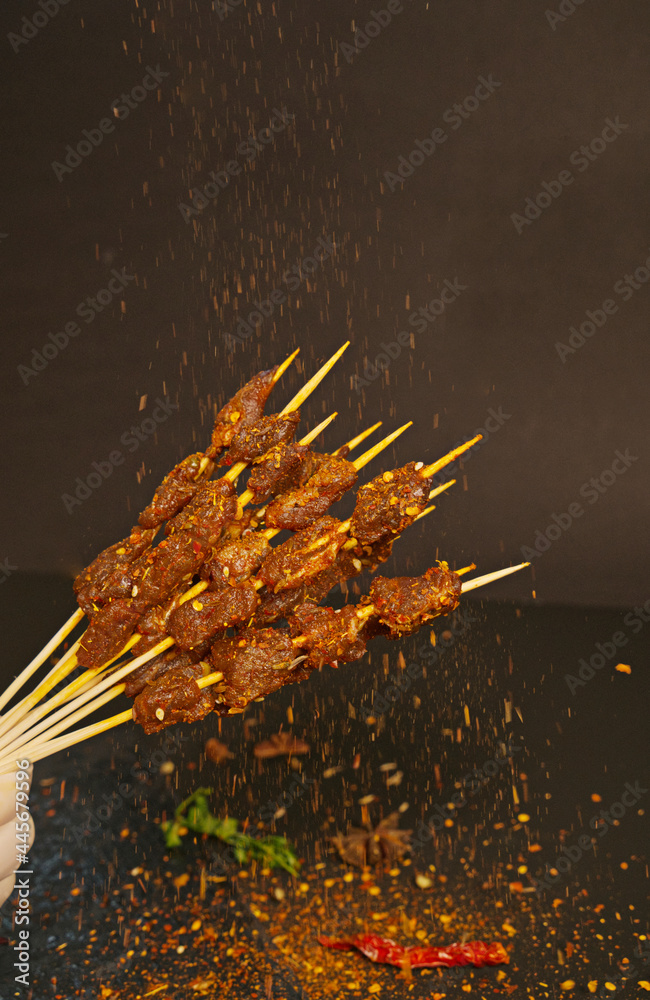 Close up of roast beef skewers in a black background picked up by a hand with seasoning sprinkled, a