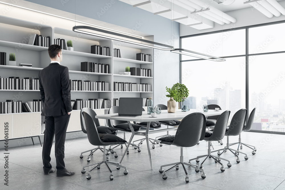 Businessman standing in modern meeting room interior with furniture, bookcase and city view. Corpora