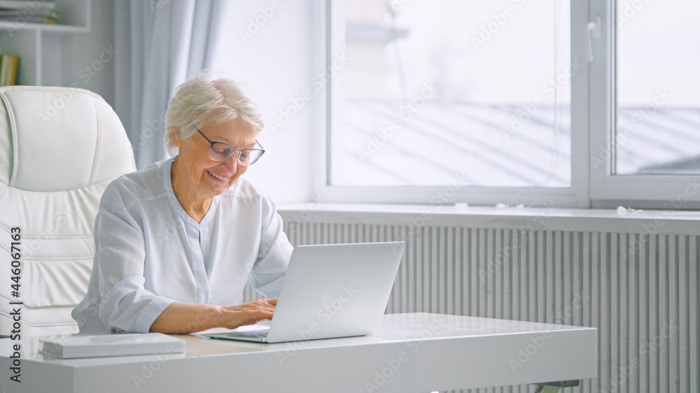 Smiling aged lady with grey hair and glasses types on laptop sitting at white table