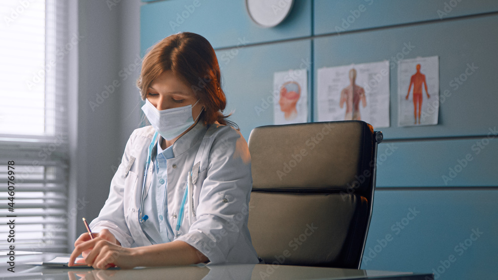 Lady general practitioner in white disposable face mask writes prescription and talks to patient