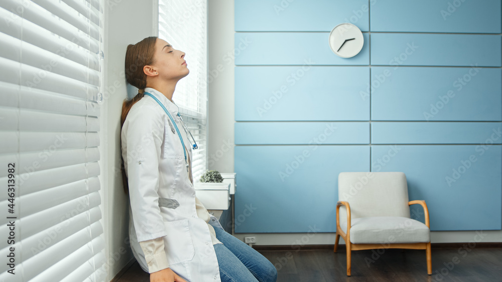 Exhausted young woman doctor in white coat sits down to rest on grey designed windowsill