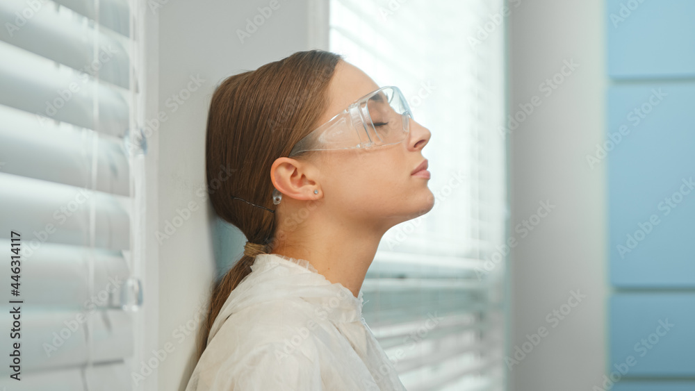 Young woman doctor in special protective coat and glasses breathes deeply leaning on wall