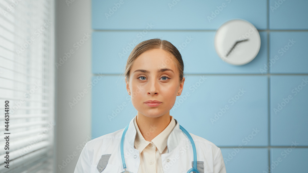 Blonde young woman general practitioner in white coat with blue stethoscope looks straight