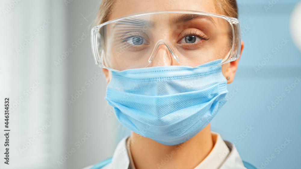 Concentrated lady doctor in blue disposable facial mask and protective goggles looks straight