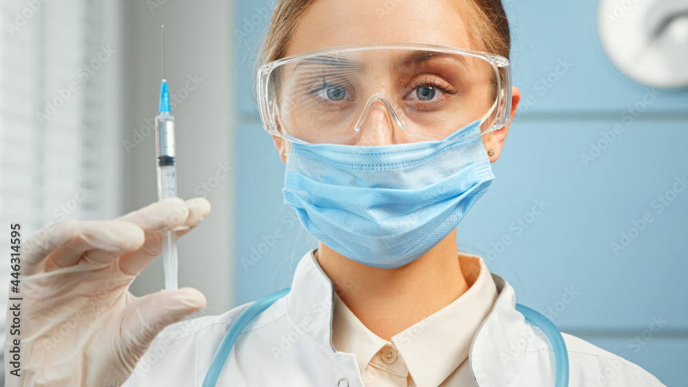 Concentrated young woman general practitioner in disposable mask and glasses holds syringe