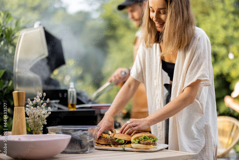 Young people making burgers at picnic with grill, woman putting cooked burgers on a plate and smile.