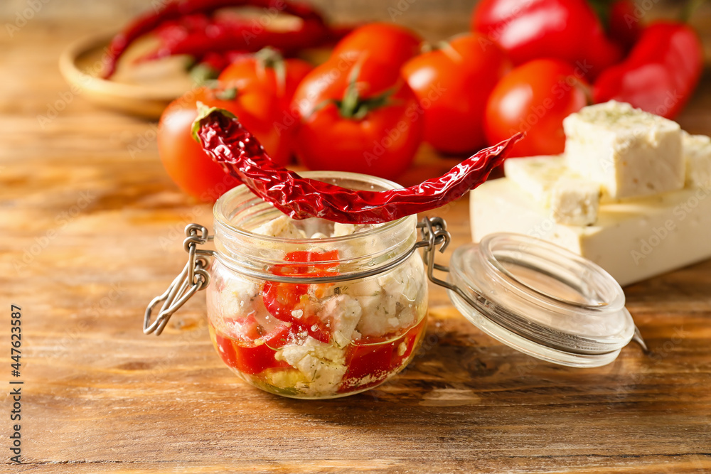 Glass jar with tasty feta cheese, oil and chili pepper on wooden background, closeup