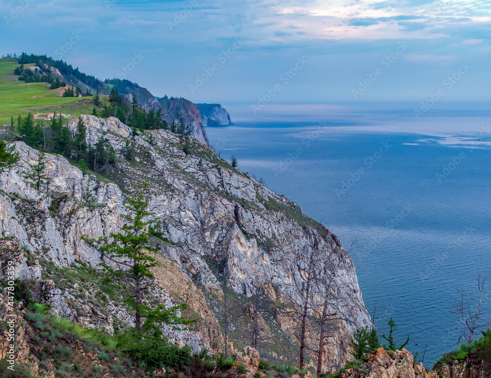 Rocky steep shore, overgrown with trees. Clouds over the water.