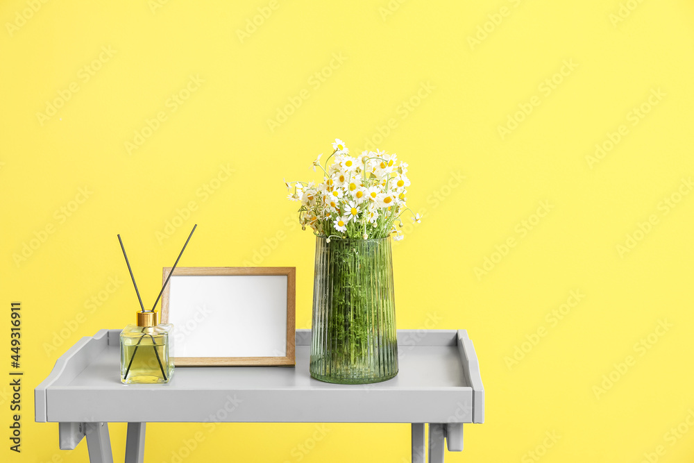 Vase with chamomiles, frame and reed diffuser on table near color wall