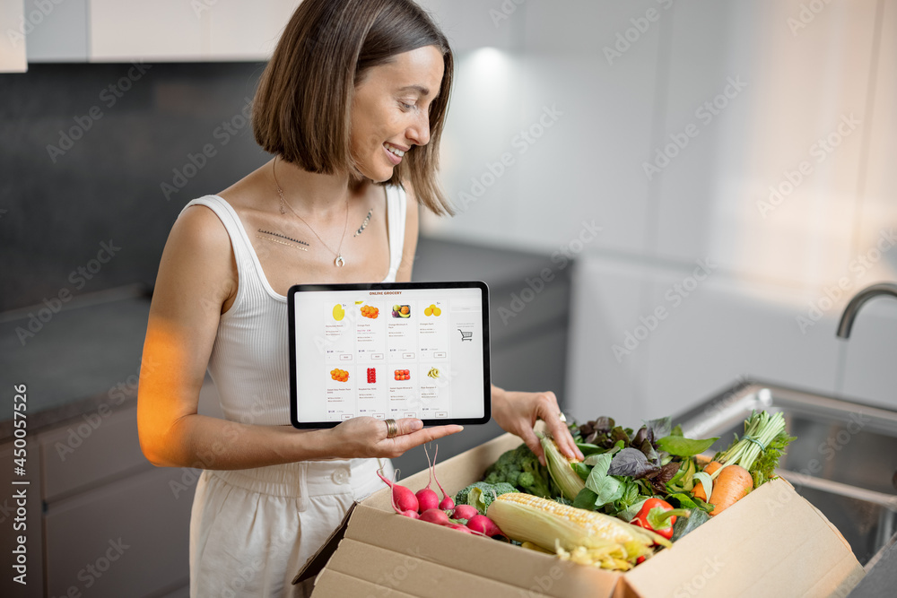 Woman buying fresh vegetables online, standing with package full of food and holding digital tablet 