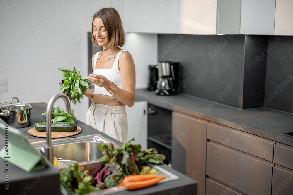 Young woman cooking with fresh vegetables and greens looking at recipe on the digital tablet at mode