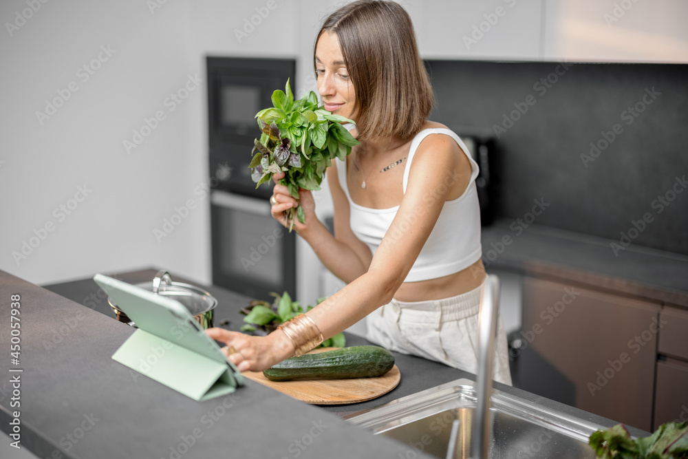 Young woman cooking with fresh vegetables and greens looking at recipe on the digital tablet at mode