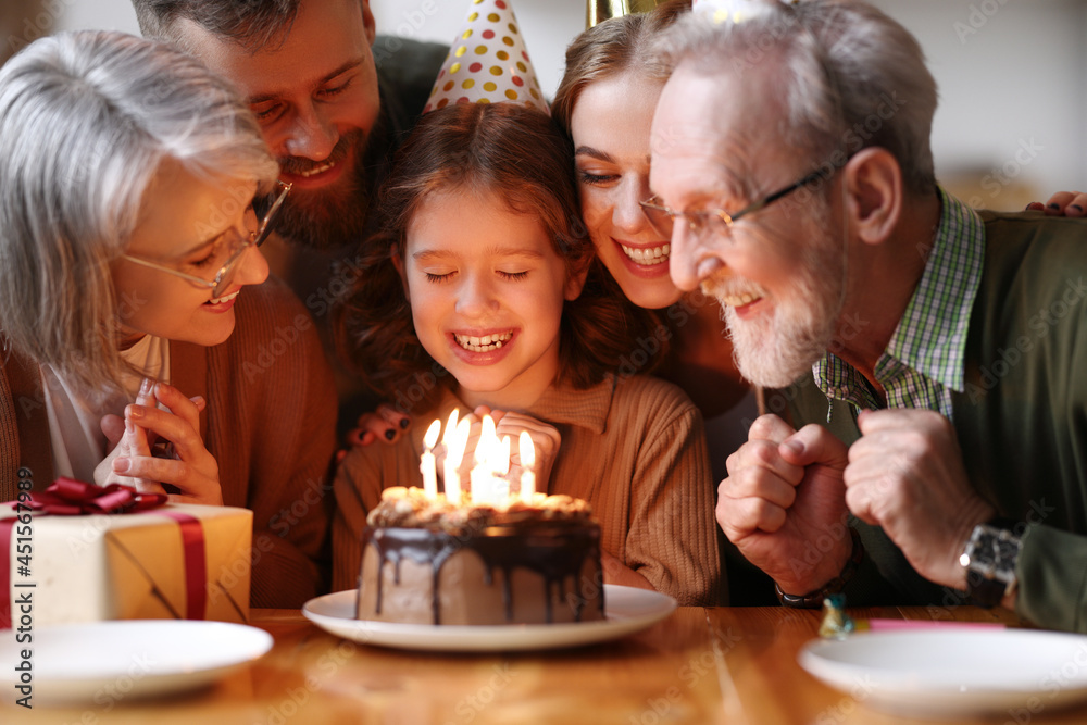 Cute excited little child making wish, blowing candles on cake while celebrating Birthday with famil