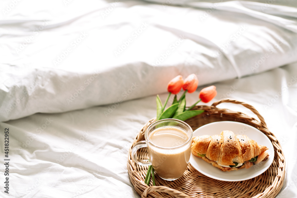 Croissant and coffee on white bed on wicker tray with flowers