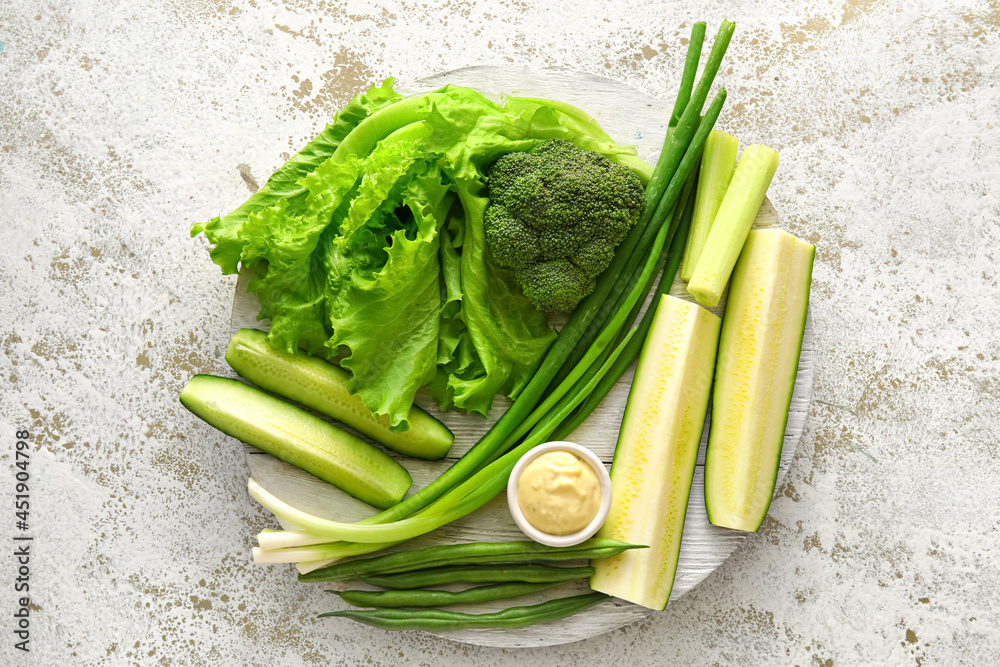 Board with different green vegetables and bowl with sauce on light background