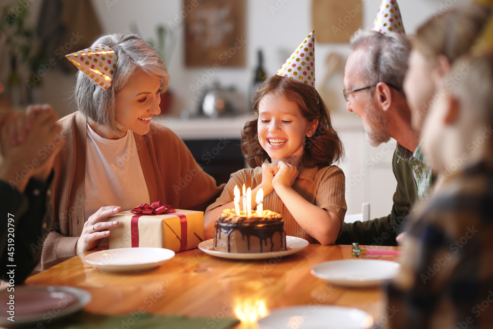 Happy little girl celebrating birthday with family at home, looking at cake with lit candles