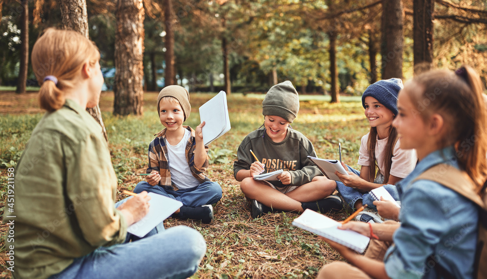 School children with notebooks and female teacher sitting on ground in forest during ecology lesson