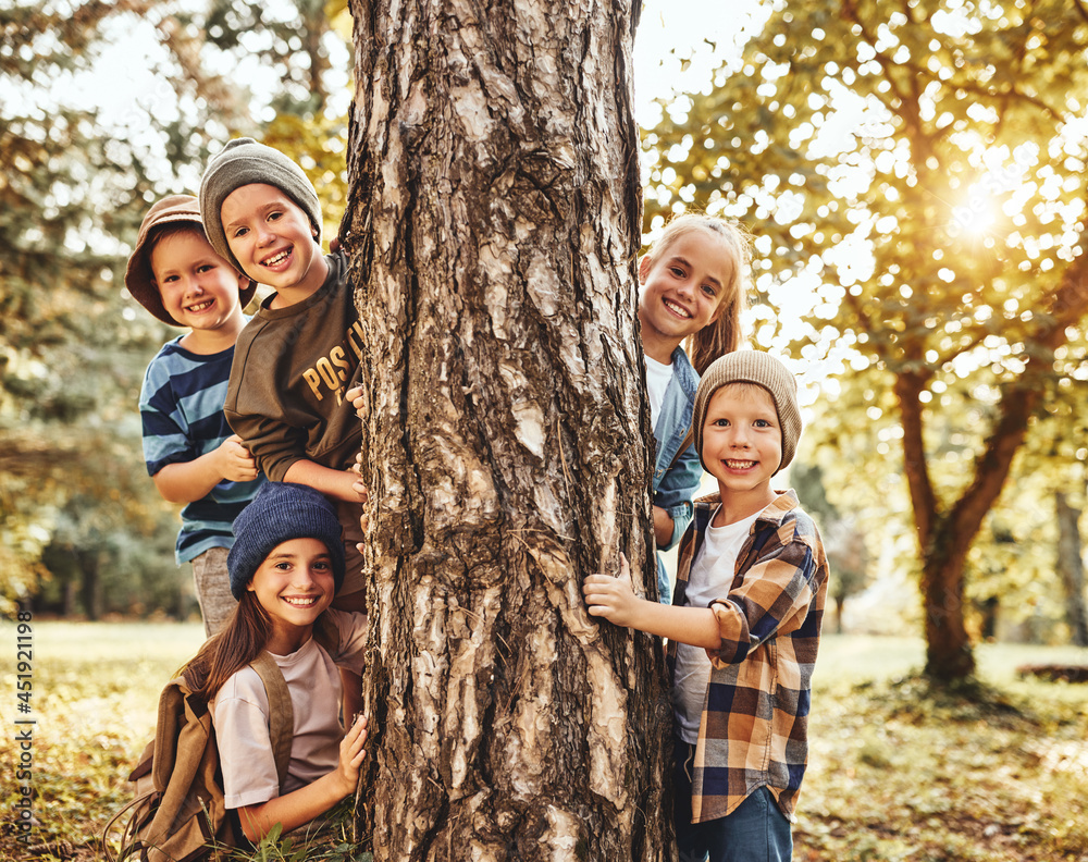 Happy smiling school children boys and girls in casual clothes playing near tree in sunny forest