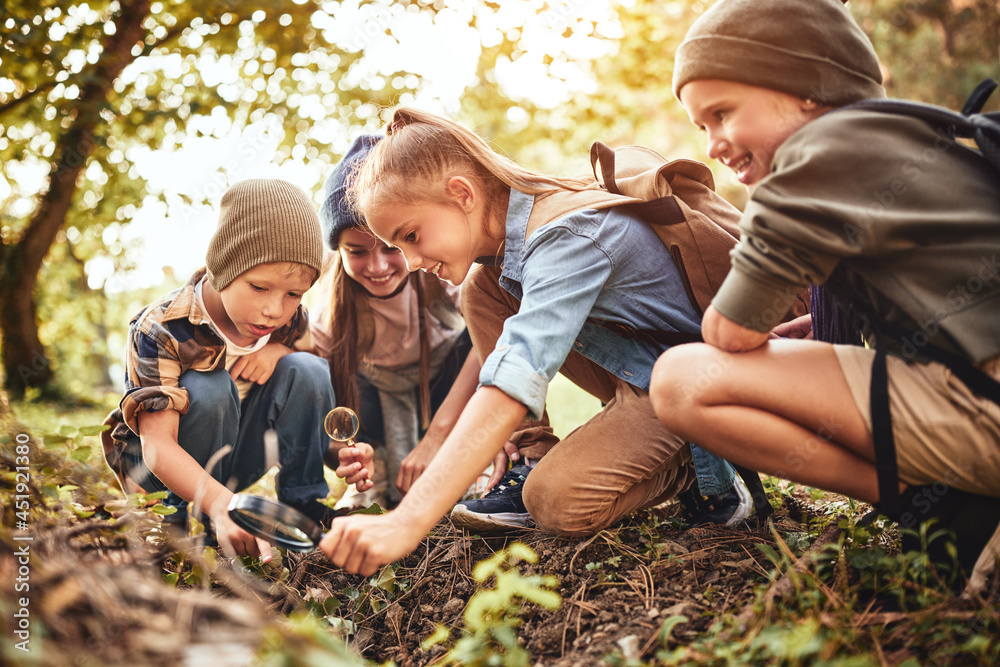 Happy school children boys and girls with backpacks making bonfire with magnifying glass in forest