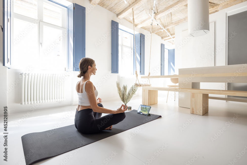 Young woman meditating and practicing yoga in a modern bright living room at home. Sports, feeling w