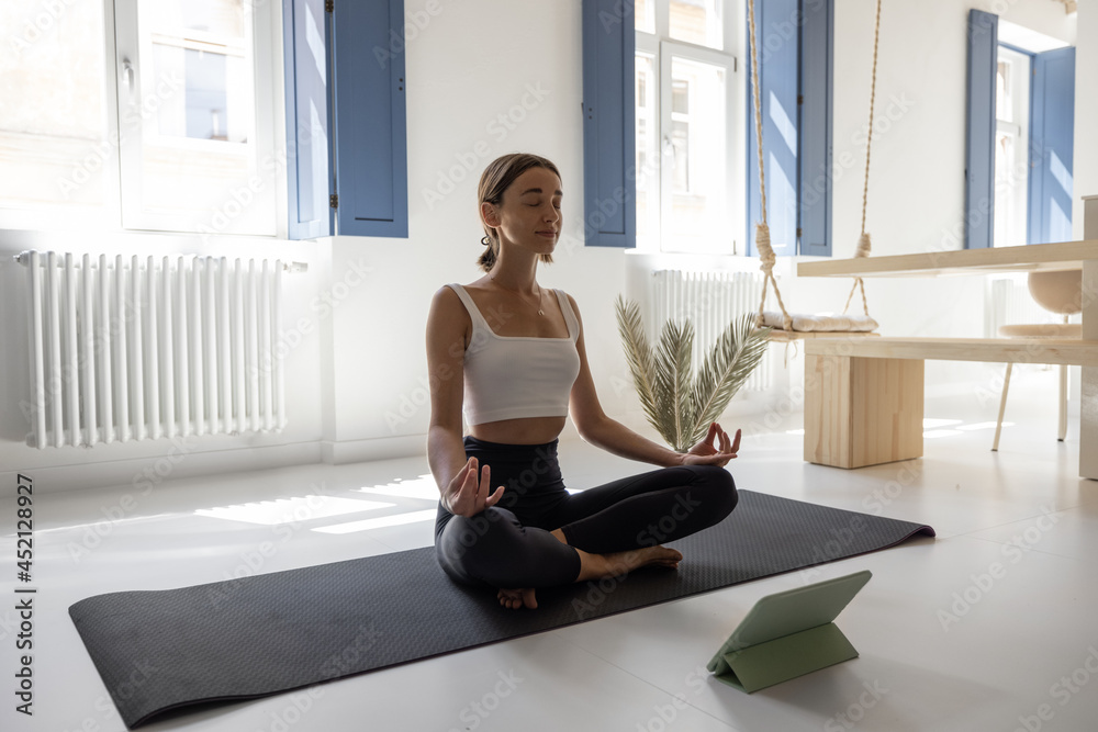 Young woman meditating and practicing yoga in a modern bright living room at home. Sports, feeling w