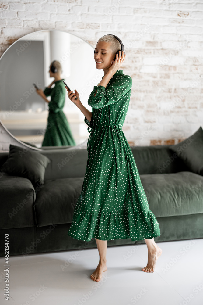 Stylish woman in green dress enjoys the music with headphones and cell phone dancing in the living r