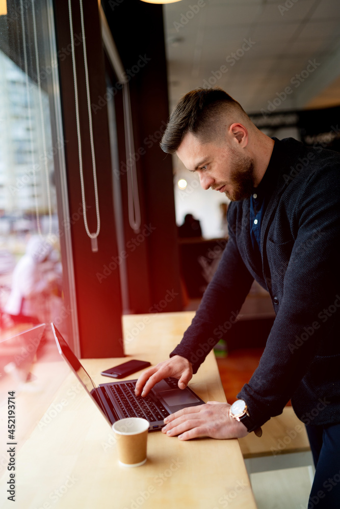 Young businessman drinking coffee and working on laptop in a cafe. Handsome man in dark suit standin