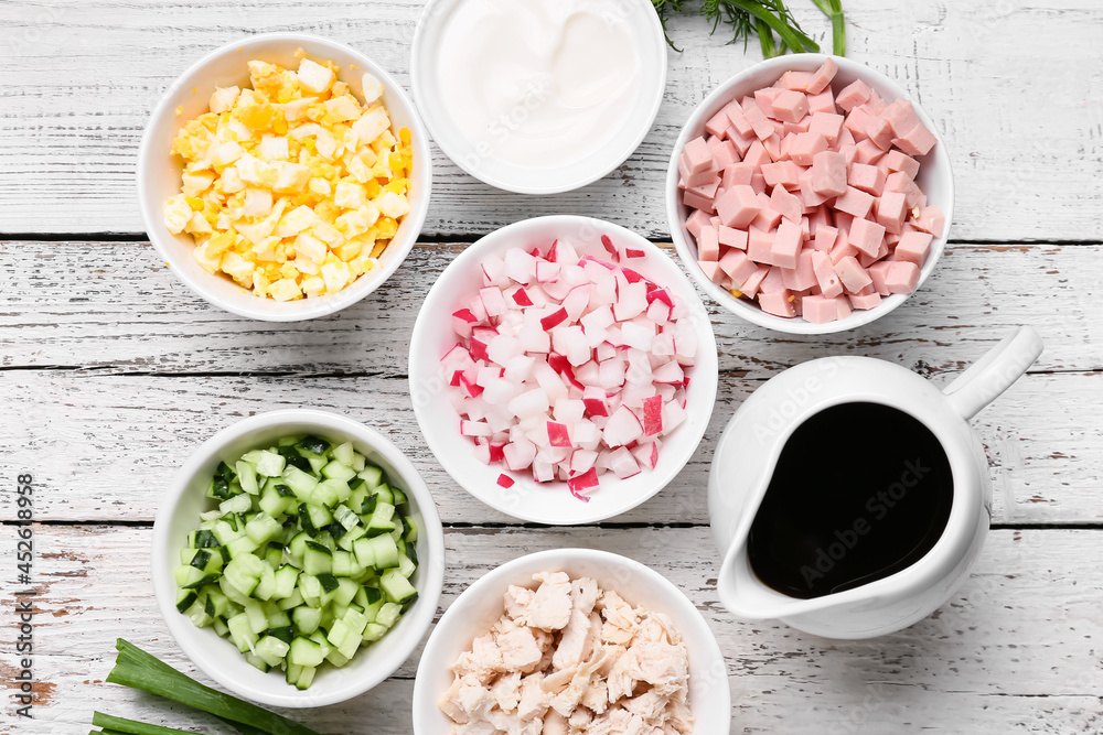 Bowls with different ingredients for okroshka and jug with kvass on light wooden background