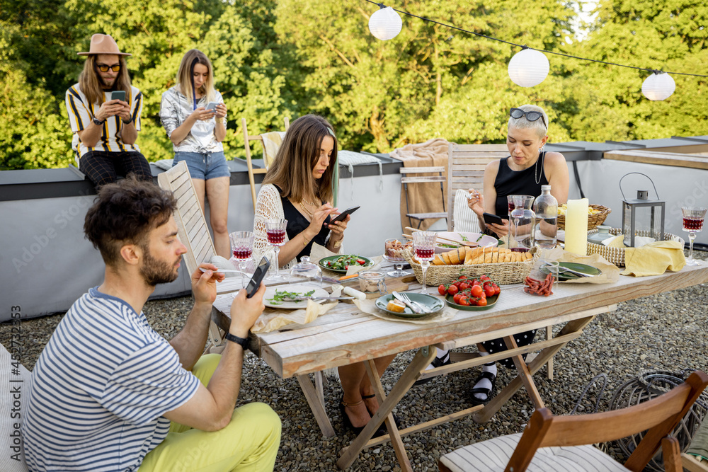 Young people sitting in smartphones separately, sadly spending time at a party on a rooftop terrace.