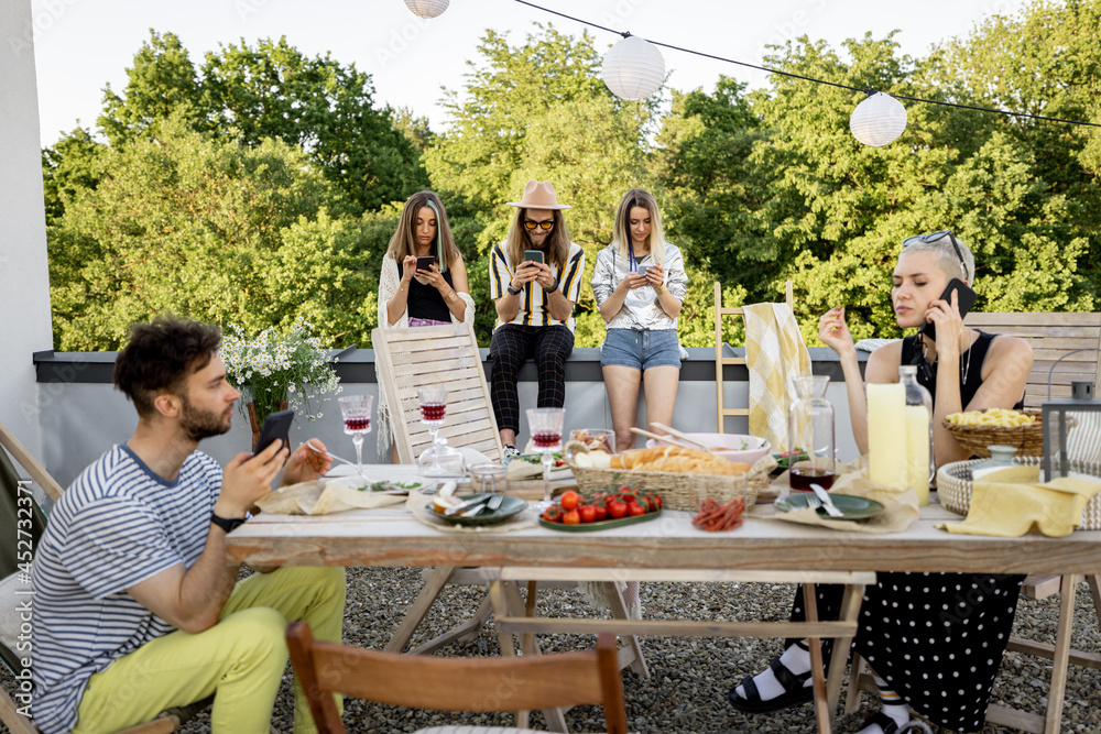 Young people sitting in smartphones separately, sadly spending time at a party on a rooftop terrace.