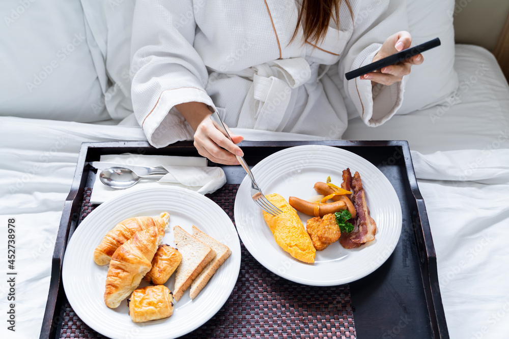 Portrait beautiful young Asian woman wearing bathrobe using smartphone while eating breakfast on a b