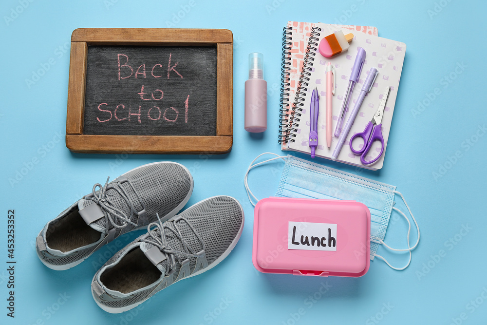 Board with text BACK TO SCHOOL, stationery, lunchbox and shoes on color background