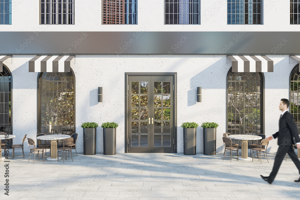 Young businessman in suit walking past creative concrete cafe exterior with terrace furniture in day