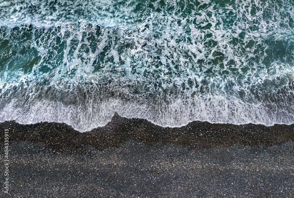 暴风雨般的绿色海浪和白色的海水泡沫在深色沙滩上破碎，从正上方观看