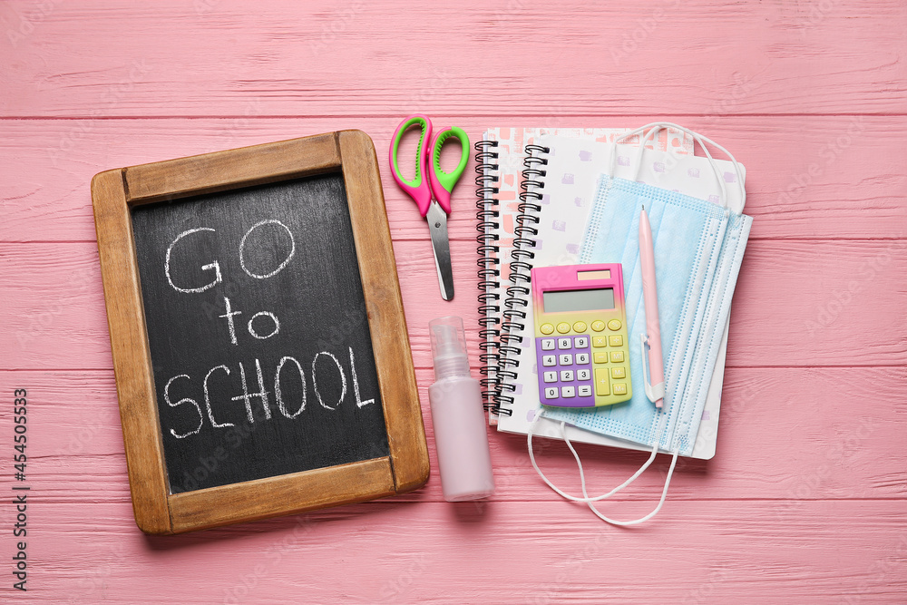 Board with text GO TO SCHOOL, stationery and medical masks on color wooden background