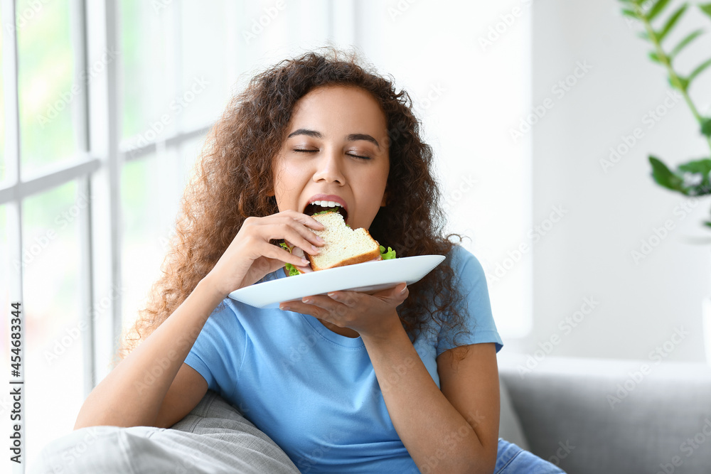 Young African-American woman eating tasty sandwich at home