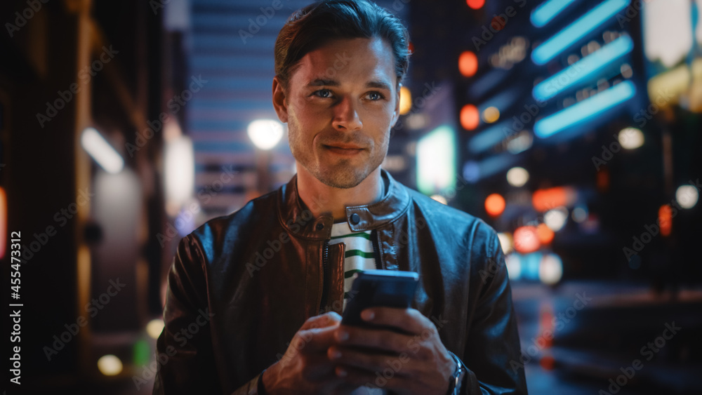 Man Using Smartphone Walking Through Night City Street Full of Neon Light. Smiling Stylish Man Using