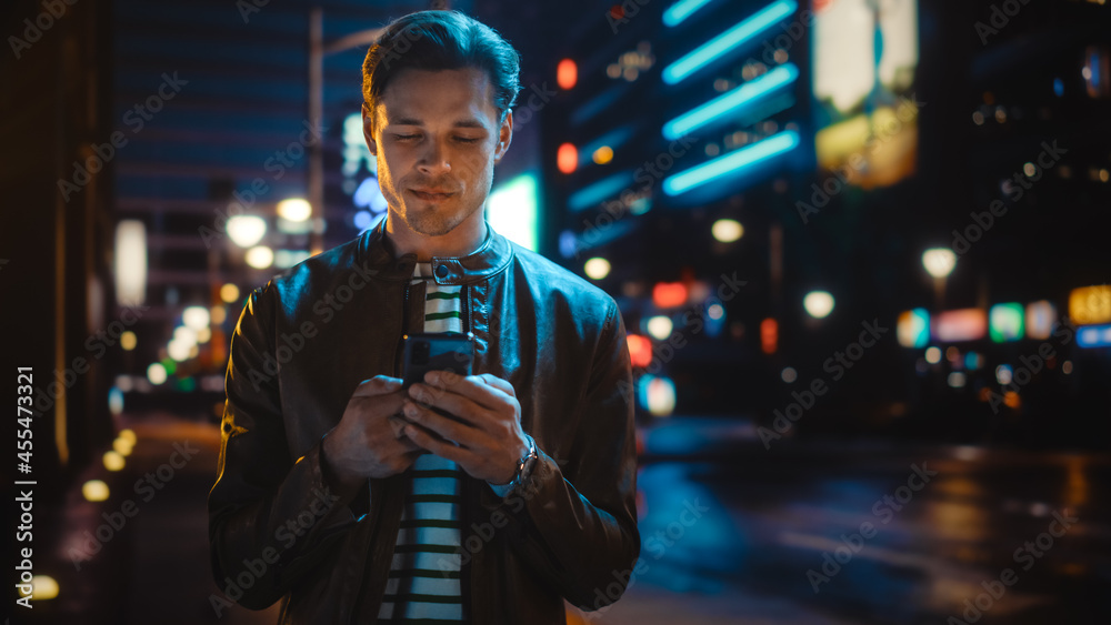 Man Using Smartphone Walking Through Night City Street Full of Neon Light. Smiling Stylish Man Using