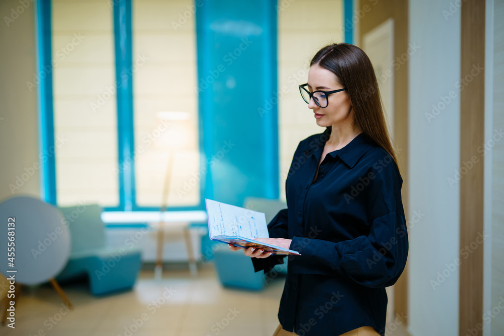 Stunning beautiful girl wearing glasses smiling and reading book in front of the camera. Light inter