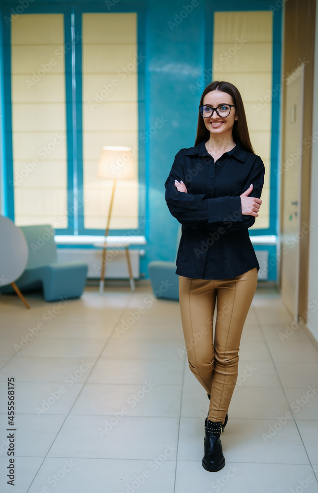 Attractive woman in dark shirt and glasses standing arms crossed in hall and smiling. Female appeara