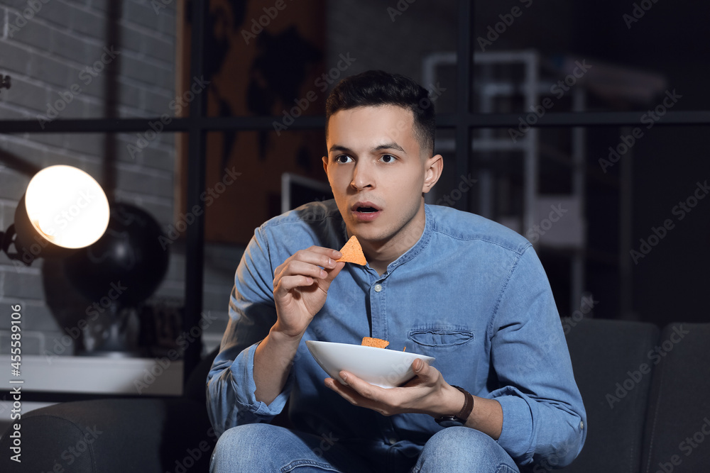 Handsome young man eating tasty nachos while watching TV at home late in evening