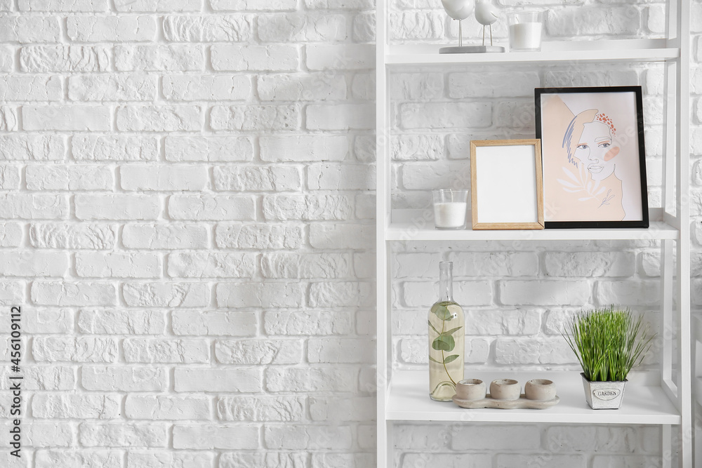 Shelf unit with modern painting, blank photo frame and different decor near white brick wall