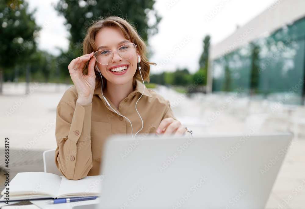 Happy young female teacher in earphones sitting in front of laptop outside having lesson online