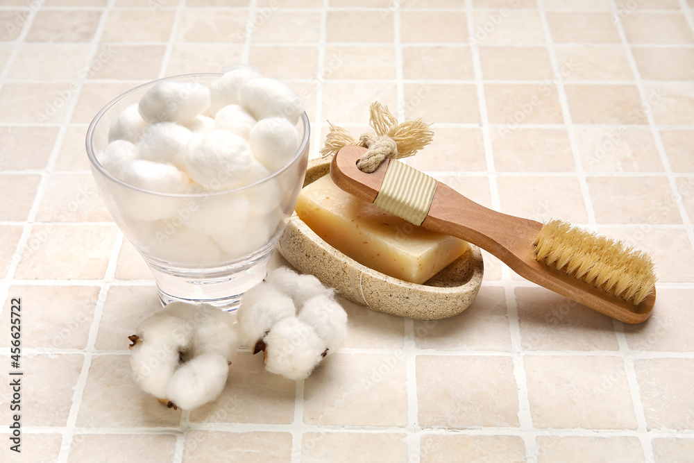 Body brush, cotton balls and soap on light tile background