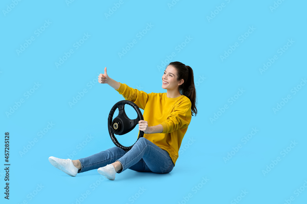 Young woman with steering wheel showing thumb-up on color background