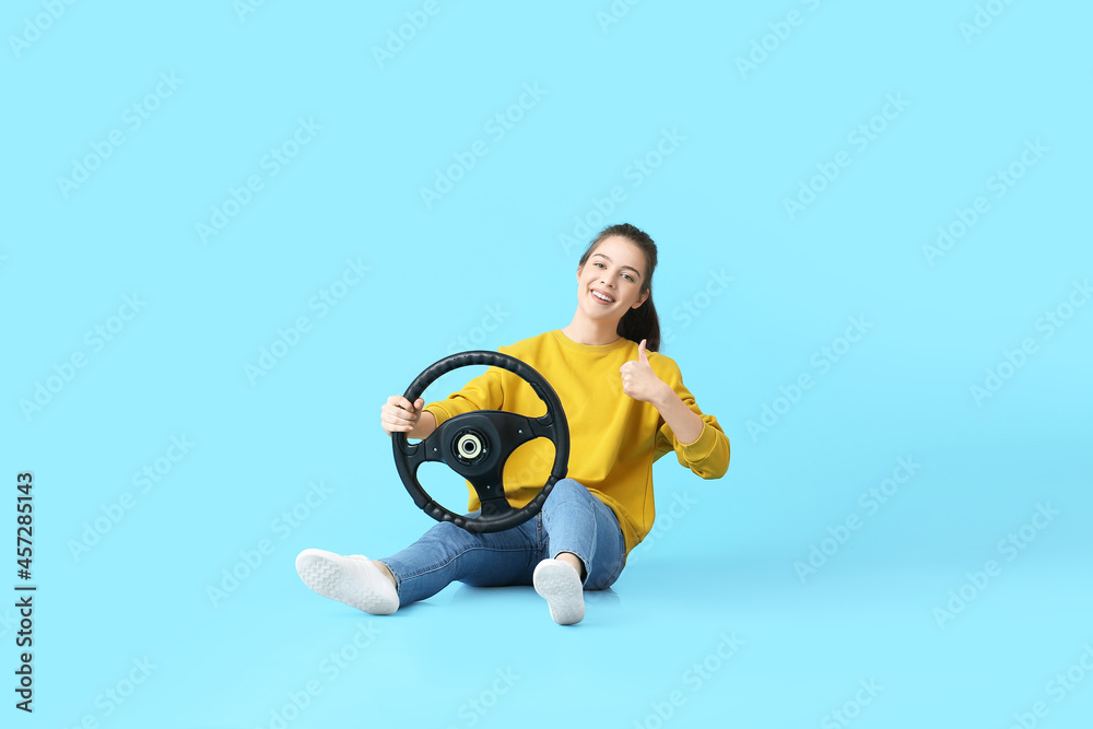 Young woman with steering wheel showing thumb-up on color background