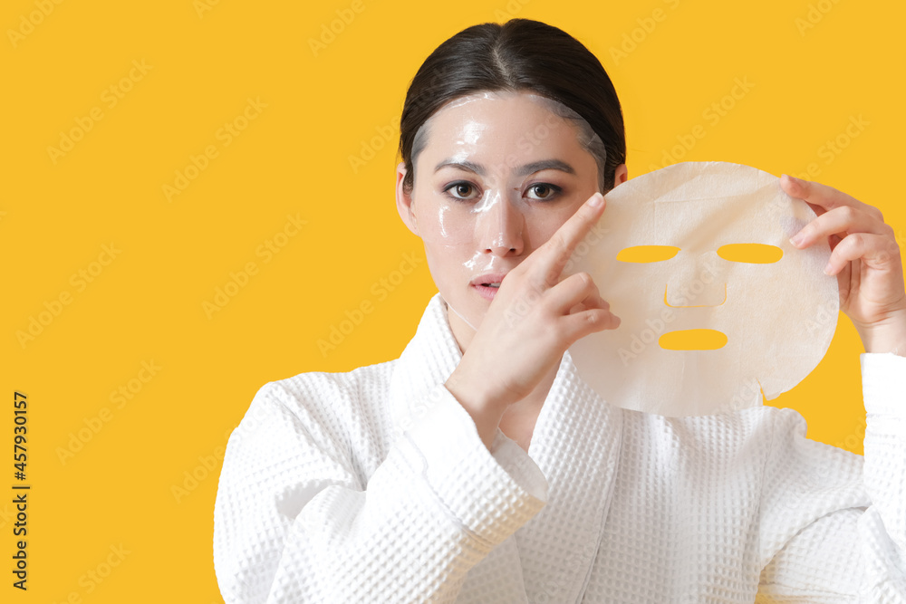 Beautiful young Asian woman with sheet facial mask on color background